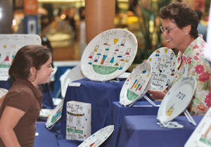 Carol of Carol Creations with customer - Carol Creations is the creator of hand-painted, custom crafted plates, platters, hot plates, mugs and more.  Carol Creations starts with high quality porcelain dinner and tableware that is then hand-painted.  After painting, each item is oven fired for durability.  Each figure is customized to resemble family members or friends with hair color and style, and other distinguishing characteristics.  Carol Creations custom painting makes these truly special and unique gifts for holidays, baby showers, weddings, birthdays . . . gifts for someone special anytime.  